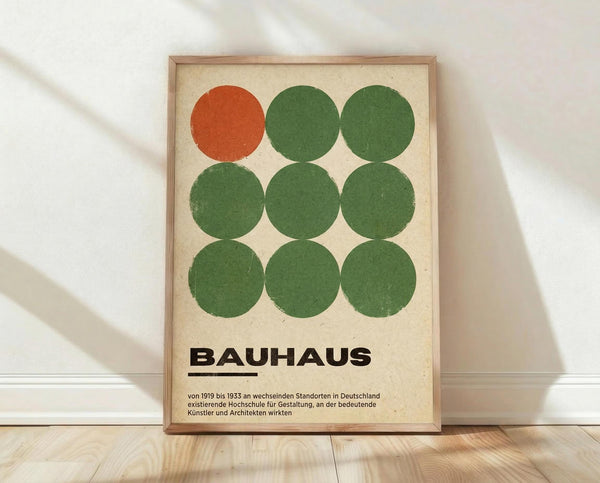 May include: Framed Bauhaus-inspired poster with nine circles arranged in a 3x3 grid - one orange circle in top left corner, eight green circles filling remaining positions. Cream-colored background with black "BAUHAUS" text and German description below. Natural wood frame, leaning against white wall on light wooden floor with soft shadows from window light.