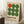 May include: Framed Bauhaus-inspired poster with nine circles arranged in a 3x3 grid - one orange circle in top left, eight green circles filling remaining positions. German text below describes the design school. Light wood frame on cream textured background, leaning against white wall corner next to white coffee cup on wooden block and beige knit throw.