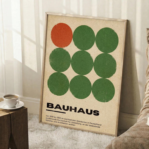 May include: Framed Bauhaus-inspired poster with nine circles arranged in a 3x3 grid - one orange circle in top left, eight green circles filling remaining positions. German text below describes the design school. Light wood frame on cream textured background, leaning against white wall corner next to white coffee cup on wooden block and beige knit throw.