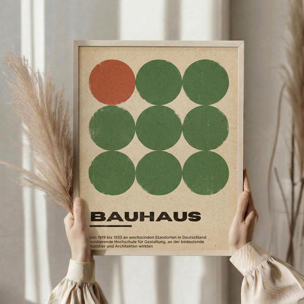 May include: Person holding framed Bauhaus-style poster featuring nine circles in a 3x3 grid - one rust red circle in top left, eight green circles filling remaining positions, on cream textured background. Black text below circles reads "BAUHAUS" with German description underneath. Natural wood frame, held against neutral curtained window with dried pampas grass visible to left.