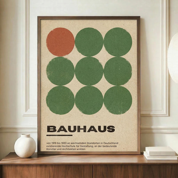 May include: Framed Bauhaus-style poster with nine circles in a 3x3 grid - one terracotta circle in top left, eight sage green circles filling remaining positions. Bold black "BAUHAUS" text below with German descriptive text underneath. Cream-colored background with brown wooden frame, displayed on wooden sideboard next to white ceramic vase and books in elegant room with white wainscoting.