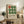 May include: Framed Bauhaus-style poster with nine circles arranged in a 3x3 grid - one orange-red circle in top left, eight green circles filling remaining positions, displayed on rustic wooden sideboard in cozy living room setting with fern plant, cream ceramic vases, books, and brown leather chair visible.