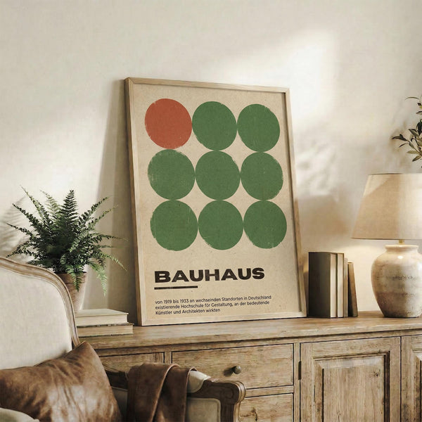 May include: Framed Bauhaus-style poster with nine circles arranged in a 3x3 grid - one orange-red circle in top left, eight green circles filling remaining positions, displayed on rustic wooden sideboard in cozy living room setting with fern plant, cream ceramic vases, books, and brown leather chair visible.