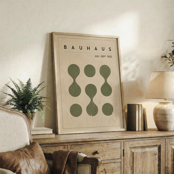 May include: Framed Bauhaus-style poster with green geometric circles arranged in a 3x3 grid pattern on beige background, displayed on rustic wooden sideboard alongside fern plant, books, ceramic vases, and brown leather chair in neutral-toned living room setting.