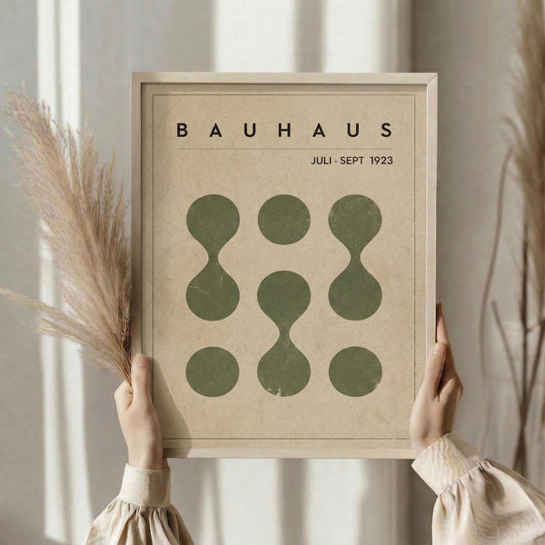May include: hands holding a framed Bauhaus-style poster with beige background and green geometric circles arranged in a 3x3 grid pattern, some forming connected shapes. Light wood frame, dried pampas grass visible to the left, soft natural lighting against cream curtained window backdrop.