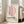 May include: Framed poster with pink curved letter D design and "BAUHAUS" text in black, displayed on light wooden floor against white wall next to beige upholstered sofa. Natural sunlight streams through tall windows creating soft shadows on the cream-colored artwork within thin wooden frame.