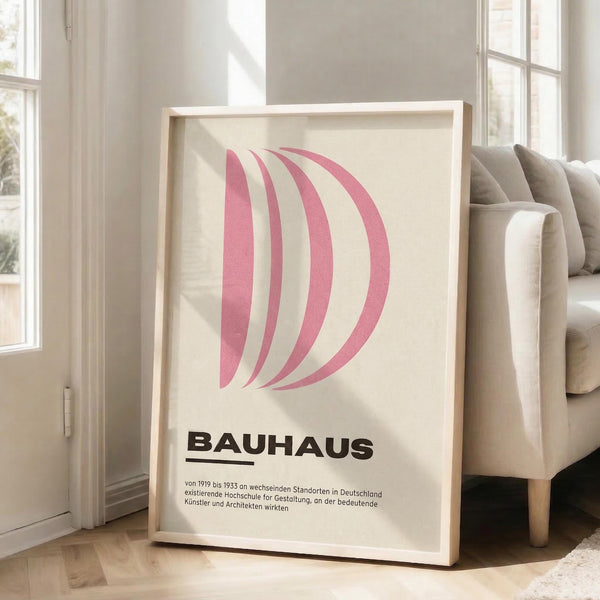 May include: Framed poster with pink curved letter D design and "BAUHAUS" text in black, displayed on light wooden floor against white wall next to beige upholstered sofa. Natural sunlight streams through tall windows creating soft shadows on the cream-colored artwork within thin wooden frame.