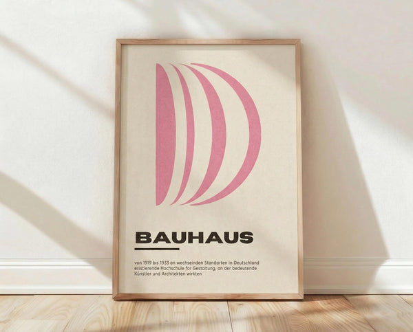 May include: Framed Bauhaus-style poster with pink curved shapes forming letter D on cream background, displayed in light wooden frame against white textured wall on wooden floor with soft natural lighting and shadows