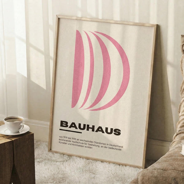 May include: Framed poster with pink curved shapes resembling the letter D on cream background, bold black "BAUHAUS" text below with German description. Light wood frame leaning against white wall on white fluffy rug. Small wooden side table with white coffee cup and saucer visible on left, beige throw blanket draped nearby. Soft natural lighting creates gentle shadows across the minimalist room setting.