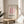 May include: Framed Bauhaus exhibition poster with pink curved geometric design displayed on light wood round side table next to beige upholstered chair. Poster features cream background with flowing pink ribbons forming abstract D shape, black text reading "BAUHAUS ausstellung" and "WEIMAR JULI-SEPT 1923". Natural sunlight creates window shadows across neutral toned room with white curtains and green plant leaves visible.