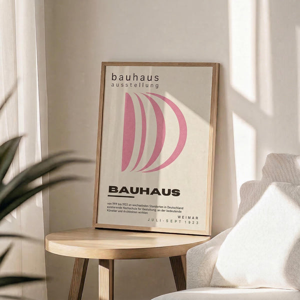 May include: Framed Bauhaus exhibition poster with pink curved geometric design displayed on light wood round side table next to beige upholstered chair. Poster features cream background with flowing pink ribbons forming abstract D shape, black text reading "BAUHAUS ausstellung" and "WEIMAR JULI-SEPT 1923". Natural sunlight creates window shadows across neutral toned room with white curtains and green plant leaves visible.