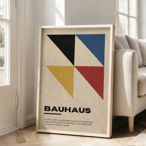 May include: Framed geometric art print with four triangular shapes in black, blue, yellow, and red on cream background, displayed against white wall next to beige upholstered chair in bright living room with large windows and natural lighting.