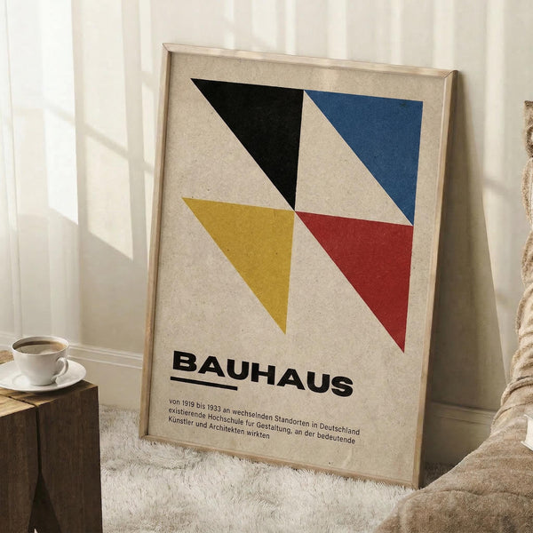 May include: Framed geometric poster with four triangular shapes in black, blue, yellow, and red on beige background, with "BAUHAUS" text and German description below. Light wood frame leaning against white wall corner beside coffee cup on wooden block and neutral textured blanket in bright natural lighting.