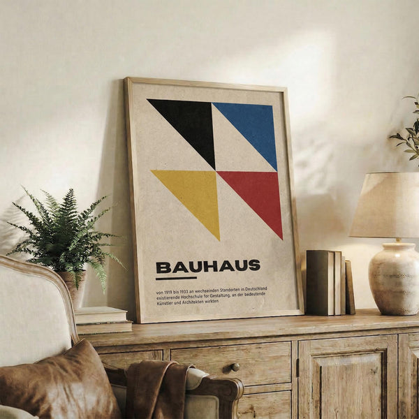 May include: Framed Bauhaus poster with geometric design featuring four colored triangles - black, blue, yellow, and red - arranged in a square pattern on cream background with German text below. Light wood frame displayed on rustic wooden sideboard alongside fern plant, ceramic vase, stacked books, and table lamp in warm-toned living room setting.