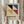 May include: Person in beige sweater holding framed Bauhaus poster featuring geometric design with black, blue, yellow, and red triangular shapes on beige background. German text below logo. Natural wooden frame, dried pampas grass visible, soft neutral lighting near window with curtains.