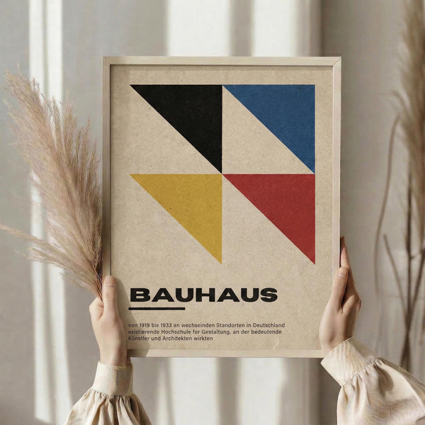 May include: Person in beige sweater holding framed Bauhaus poster featuring geometric design with black, blue, yellow, and red triangular shapes on beige background. German text below logo. Natural wooden frame, dried pampas grass visible, soft neutral lighting near window with curtains.