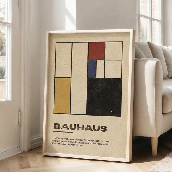 May include: Framed Bauhaus poster with geometric design featuring colored rectangles in red, blue, yellow, and black on cream background, displayed in white frame leaning against wall next to beige sofa in bright living room with hardwood floors and large windows