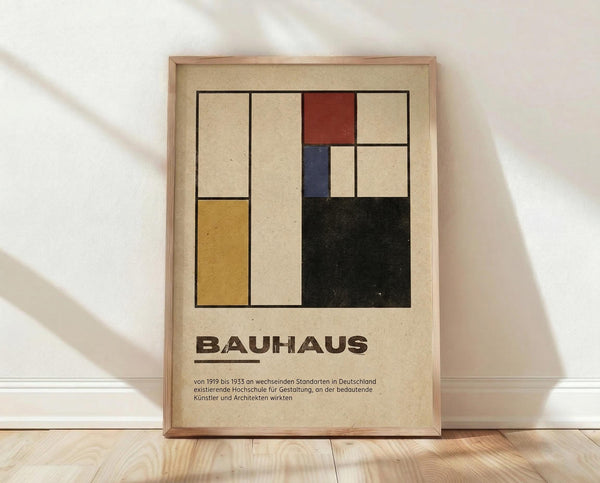May include: Framed Bauhaus poster with geometric abstract design featuring colored rectangles in yellow, red, blue, and black on cream background. German text below describes the design school from 1919-1933. Light wood frame leaning against white wall on wooden floor with soft natural lighting and fabric shadows.