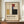 May include: Framed Bauhaus poster with geometric composition in red, blue, yellow, black and cream rectangles on beige background with German text, displayed on wooden sideboard next to white ceramic vase and stacked books in bright room with white paneled walls