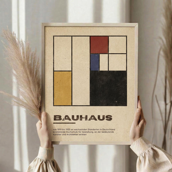 May include: Person in cream sweater holding framed Bauhaus poster with geometric rectangles in yellow, red, blue, black, and beige colors against neutral background. Dried wheat stems visible to left. Poster features German text about the design school from 1919-1933. Natural lighting creates soft shadows on white curtained backdrop.