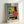 Framed abstract geometric art print in Bauhaus style featuring bold shapes in teal, yellow, orange, and black. The artwork displays architectural elements including vertical lines, circles, and angular forms arranged in a modernist composition. Text reads "BAUHAUS" vertically and "FORM FOLLOWS FUNCTION" at bottom. The print sits in a light wood frame against white walls, showing the cream-colored paper texture and "01" numbering in the corner.