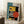May include: Framed geometric abstract poster in Bauhaus style featuring bold shapes in yellow, orange, teal, black and white with "BAUHAUS 01" and "FORM FOLLOWS FUNCTION" text, displayed leaning against white wall on fluffy white rug next to wooden side table with coffee cup and cozy chair corner in bright room with natural window light