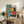 May include: Framed geometric abstract art print in Bauhaus style featuring teal, yellow, coral, black and white shapes including circles, rectangles and stripes, displayed on rustic wooden sideboard alongside books, ceramic vases, and fern plant in modern neutral living room setting