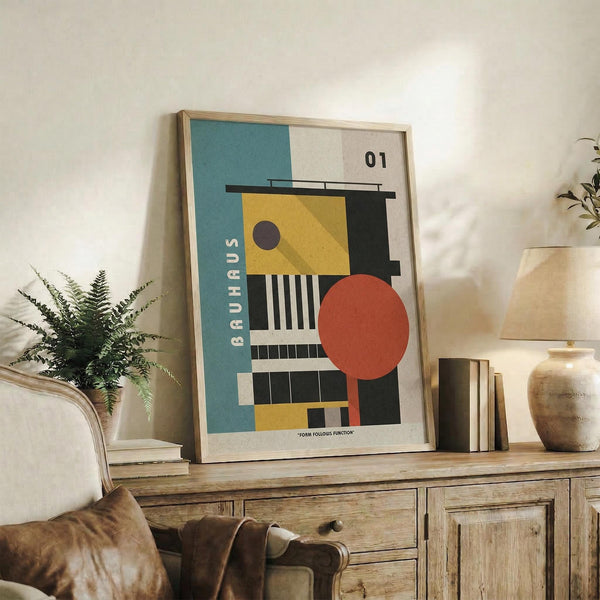 May include: Framed geometric abstract art print in Bauhaus style featuring teal, yellow, coral, black and white shapes including circles, rectangles and stripes, displayed on rustic wooden sideboard alongside books, ceramic vases, and fern plant in modern neutral living room setting