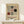 May include: Framed Bauhaus-style poster with geometric shapes in black, red, and gray on cream background, displayed on light wood console table next to small white gift box with evergreen sprig, against white wall