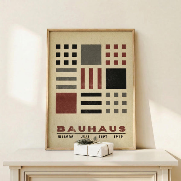 May include: Framed Bauhaus-style poster with geometric shapes in black, red, and gray on cream background, displayed on light wood console table next to small white gift box with evergreen sprig, against white wall