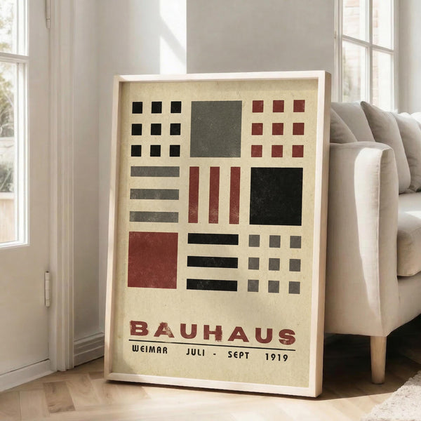 May include: Framed Bauhaus-style poster with geometric grid pattern in black, red, and gray rectangles and squares on cream background, displayed in white frame leaning against wall next to beige sofa in bright modern living room with large windows and hardwood floors.