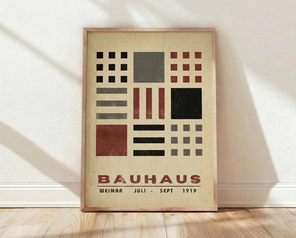 May include: Framed Bauhaus poster with geometric shapes in black, red, and gray on cream background, leaning against white wall on light wood floor. Features grid patterns, squares, rectangles, and lines in modernist style with "BAUHAUS WEIMAR JULI-SEPT 1919" text at bottom in thin wooden frame.