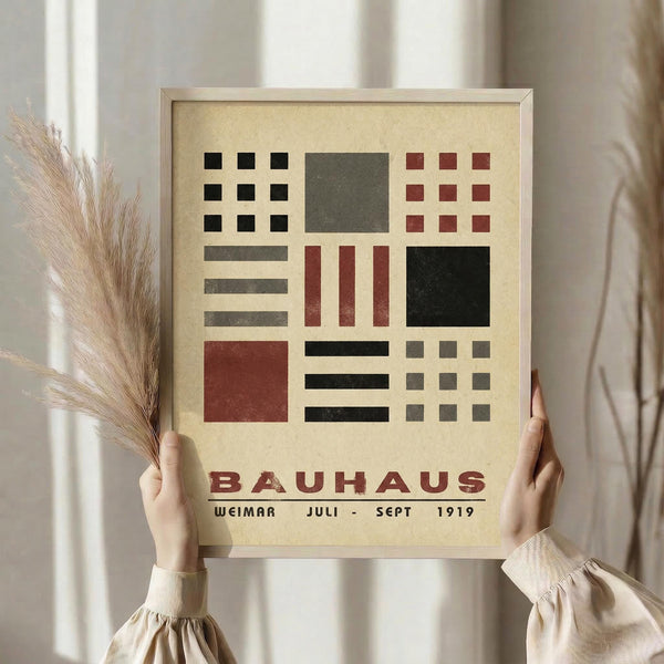 May include: Person in cream-colored clothing holding framed Bauhaus-style poster featuring geometric shapes in black, red, and gray on beige background. Dried pampas grass visible to left. Light wooden frame against neutral interior setting with soft natural lighting and beige curtains.