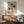 May include: Bauhaus art poster in brown frame hanging on white wall above wooden sideboard with white table lamp. Green upholstered armchair with wooden frame sits nearby with round coffee table. Sunlight casts shadows across the mid-century modern room setting. The vintage-style poster features geometric shapes in black, red, and green arranged in grid pattern on cream background.