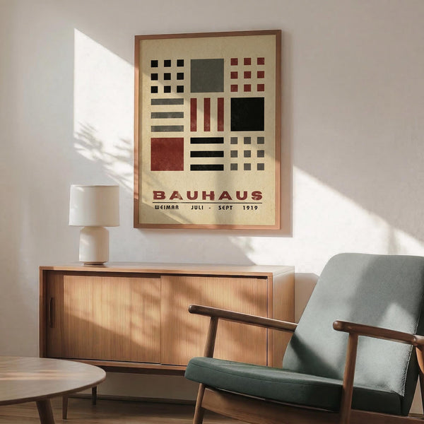 May include: Bauhaus art poster in brown frame hanging on white wall above wooden sideboard with white table lamp. Green upholstered armchair with wooden frame sits nearby with round coffee table. Sunlight casts shadows across the mid-century modern room setting. The vintage-style poster features geometric shapes in black, red, and green arranged in grid pattern on cream background.
