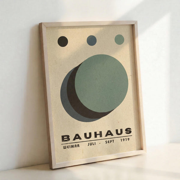 Framed vintage-style poster with geometric design featuring overlapping circles in sage green, dark green, and charcoal creating a crescent moon effect. Three small circles in matching colors appear at the top. Bold black text reads "BAUHAUS" with "WEIMAR JULI - SEPT 1919" below on cream textured background. Light wood frame sits against white wall with natural lighting creating subtle shadows.