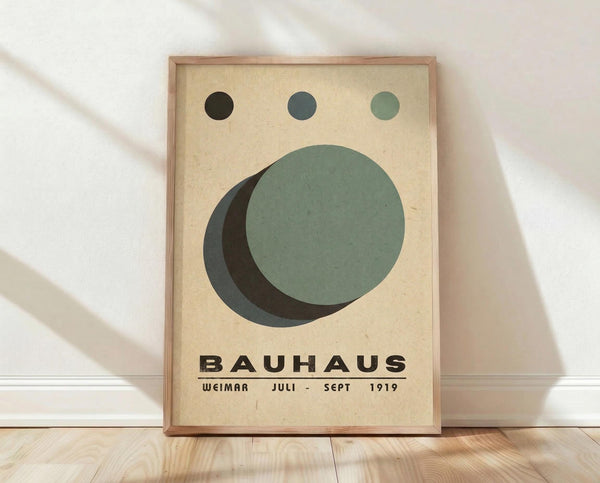 May include: Framed Bauhaus-style poster with geometric design featuring overlapping circles in sage green, dark green, and black on cream background. Three small circles aligned at top in gradient from black to light green. Bold black text reads "BAUHAUS WEIMAR JULI - SEPT 1919" at bottom. Light wood frame sits on wooden floor against white wall with soft natural lighting and shadows.