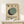 May include: Framed Bauhaus-style poster with geometric design featuring overlapping circles in sage green, dark green, and charcoal gray on cream textured background, displayed on light wood console table against white wall with small wrapped gift box beside it