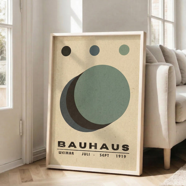 May include: Framed Bauhaus-style art print leaning against white wall next to beige sofa. Poster features cream background with geometric shapes - three small circles in black, teal, and sage green at top, large overlapping circles in sage green and dark teal below, black text reading "BAUHAUS" and "WEIMAR JULI-SEPT 1919" at bottom. Natural wood frame, bright room with large windows.