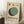 May include: Framed Bauhaus-inspired poster with geometric design featuring overlapping circles in sage green, dark green, and navy blue on cream textured background, displayed in light wooden frame leaning against white wall corner, with coffee cup on wooden block and beige textile nearby on fluffy white rug.
