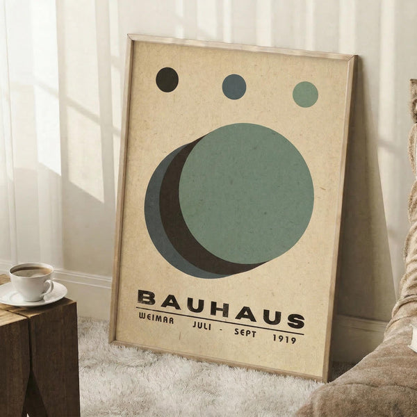 May include: Framed Bauhaus-inspired poster with geometric design featuring overlapping circles in sage green, dark green, and navy blue on cream textured background, displayed in light wooden frame leaning against white wall corner, with coffee cup on wooden block and beige textile nearby on fluffy white rug.