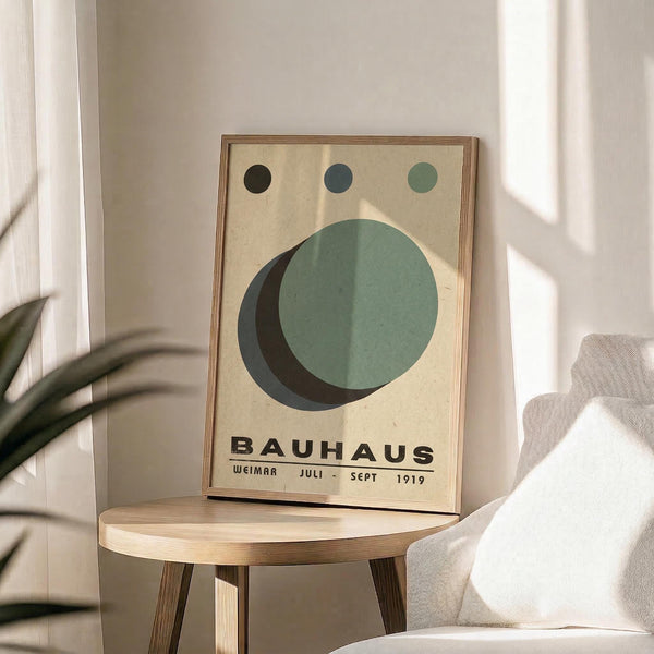 May include: Framed Bauhaus-style poster with geometric design featuring large overlapping circles in sage green and black, three small circles above in gradient from black to green, cream background, displayed on round wooden side table next to beige upholstered chair with plant leaves visible in corner, natural lighting creating soft shadows on white wall
