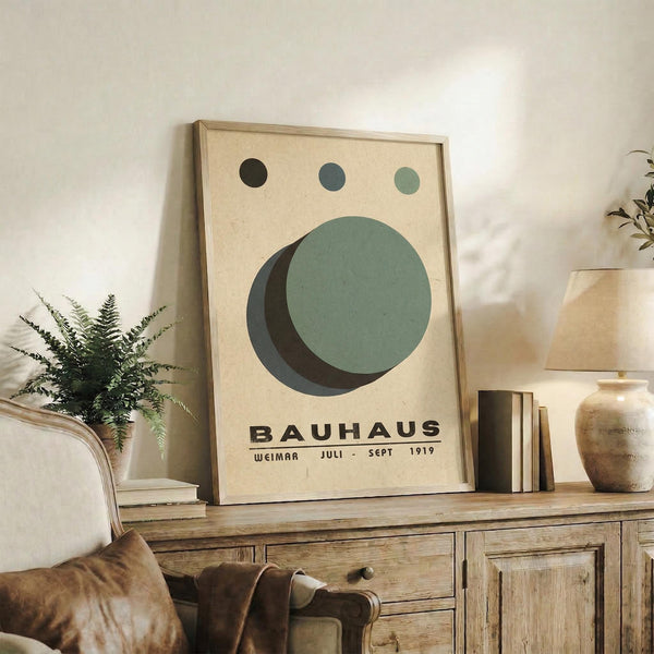 May include: Framed Bauhaus-style poster with geometric circles in sage green, dark green, and black on cream background, displayed on rustic wooden sideboard alongside books, ceramic vase, and potted fern in modern living room setting with leather armchair and warm neutral tones.