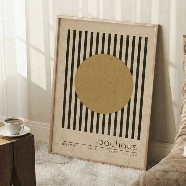 May include: Framed Bauhaus-style poster with beige textured background, black vertical lines, and tan circle design element. Light wood frame leaning against white wall corner next to white coffee cup on wooden block and beige knit throw blanket on fluffy white rug.