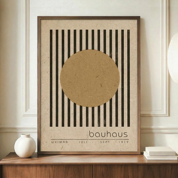 May include: Minimalist Bauhaus-style poster print in wooden frame on wood credenza. Features vertical black lines with overlapping tan circle on cream textured background. Text reads "bauhaus" and "weimar juli - sept 1919" in lowercase letters. Displayed alongside white ceramic vase and stacked books against cream walls with white molding in modern interior setting.