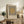May include: Framed Bauhaus-style poster with black vertical stripes and tan circle on cream background, displayed on rustic wooden sideboard beside fern plant, books, ceramic vase, and brown leather chair in neutral-toned living room setting with natural lighting