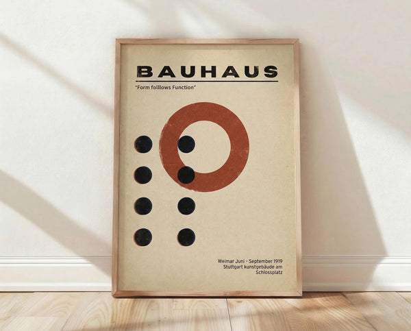 May include: Framed Bauhaus-style poster with cream background featuring bold black "BAUHAUS" text, red-orange circle, and geometric black dots arrangement. Light wood frame leaning against white wall on natural wood floor with soft window light creating shadows.