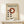 May include: Framed Bauhaus poster with cream background featuring red circle and black dots in geometric arrangement, displayed on light wood shelf next to small wrapped gift with twine and greenery sprig against white wall