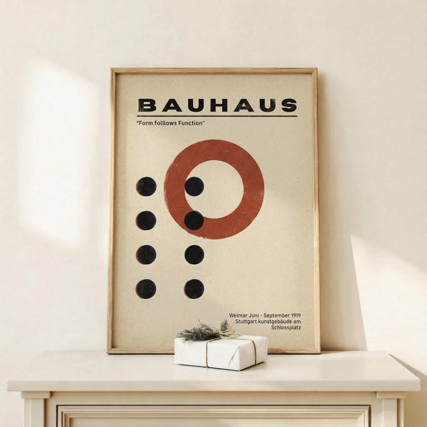May include: Framed Bauhaus poster with cream background featuring red circle and black dots in geometric arrangement, displayed on light wood shelf next to small wrapped gift with twine and greenery sprig against white wall