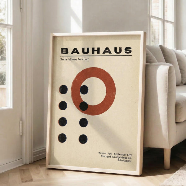 May include: Framed Bauhaus-style poster with cream background featuring red circle and black dots in geometric arrangement, displayed against white wall next to beige sofa in bright living room with large windows and natural light streaming across wooden floor